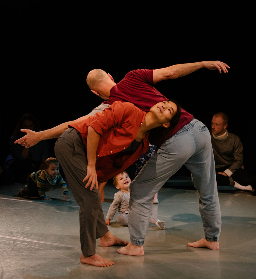 Tow dancers interact with eachother back-to-back, with babies and toddlers watching them from the side