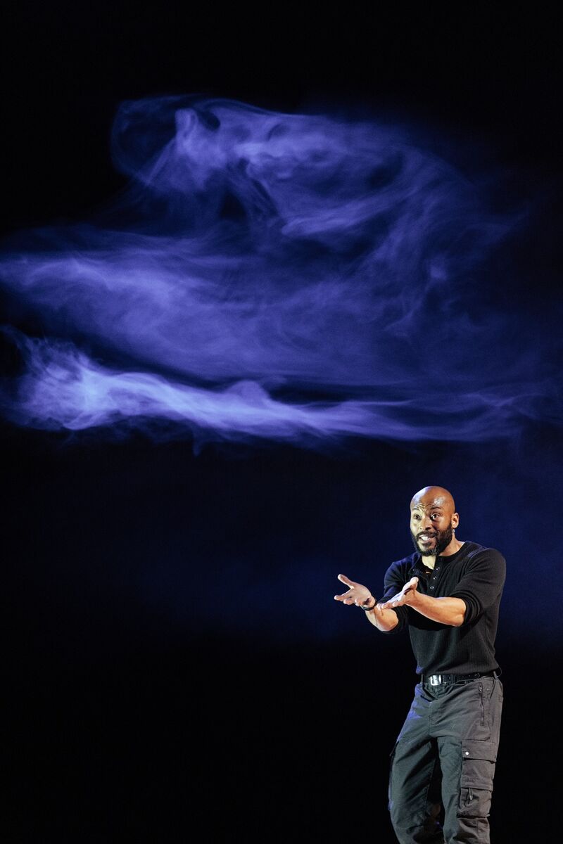 Arinze, a black man wearing an all black outfit, stands with hands raised, as if he is pleading with someone. Above him is a blue smoky cloud