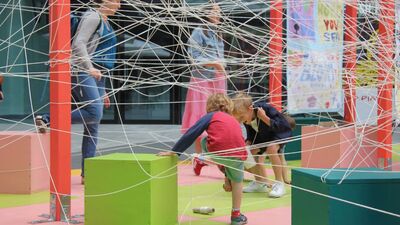 Two young children engage with an installation made of brightly coloured columns and boxes criss crossed with lots of string