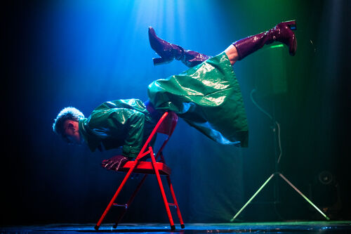 An artist performs chair acrobatics on a bright red chair. They are dressed in a green PVC coat and knee-high, burgundy PVC boots.