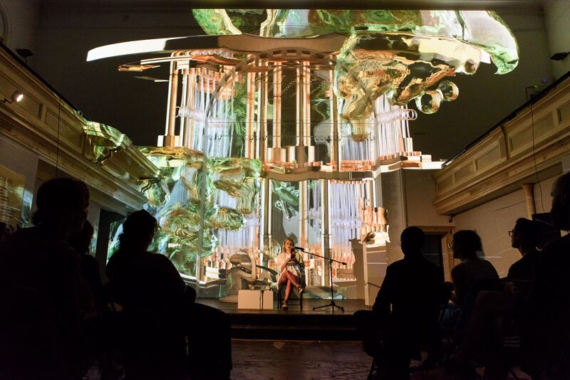 Libby Heaney, a white woman, sits on stage. There are large projections of her artwork behind her and an audience is silhoutted in front of her