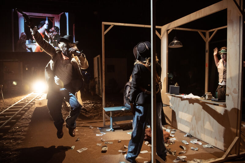 Actors dressed as cowboys perform a dramatic scene in a stage set resembling a bank, with fake money scattered on the floor and dynamic lighting highlighting the action.