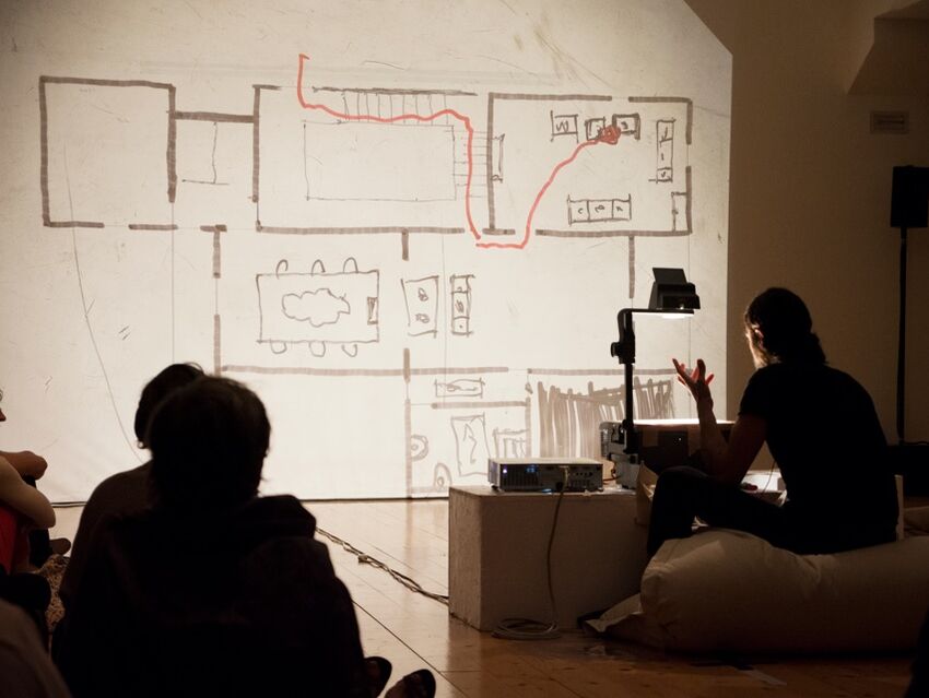 Photograph showing a projection on a white wall with a performer speaking. The projection shows the floor plan to a building with a red line drawn through it to show where someone might walk