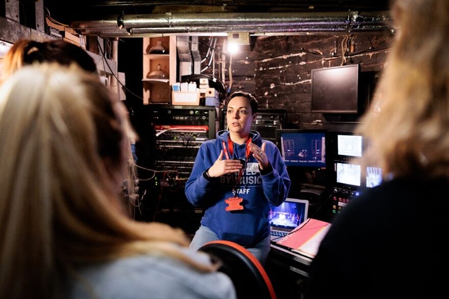 A woman standing backstage at a theatre talks to a group of people