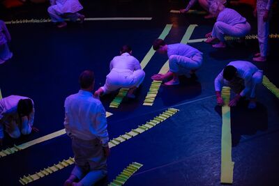 A collection of people dressed in white jumpsuits are squating on the floor, ruffling up long strips of yellow paper
