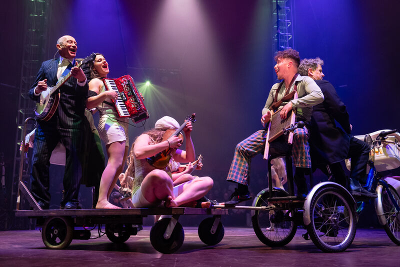 A group of musicians playing a banjo, accordian, violin are moving around on a wheeled platform pulled by a big trike