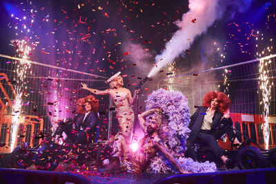 A performance shot from Mr. Blackpool. Four performers are in various seated and standing positions centre stage, with confetti and extravagant outfits. 