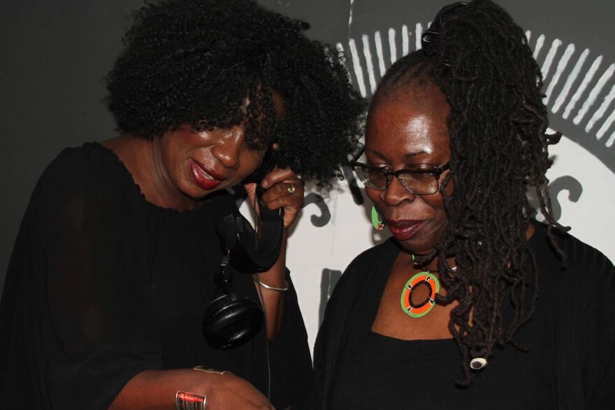 Two black women standing nexdt to each other, one is holdng a headphone up to her ear