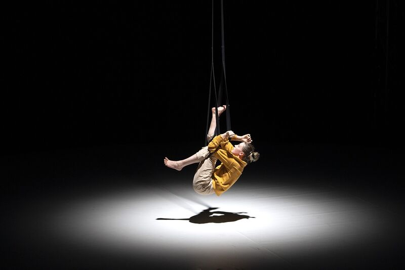 In a spotlight, a woman in a yellow jacket is suspended just off the floor, holding on to some black ropes