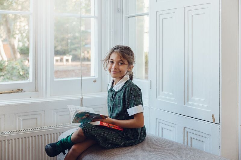 A young school girl sitting on a bench in a white-walled room, she is reading a book