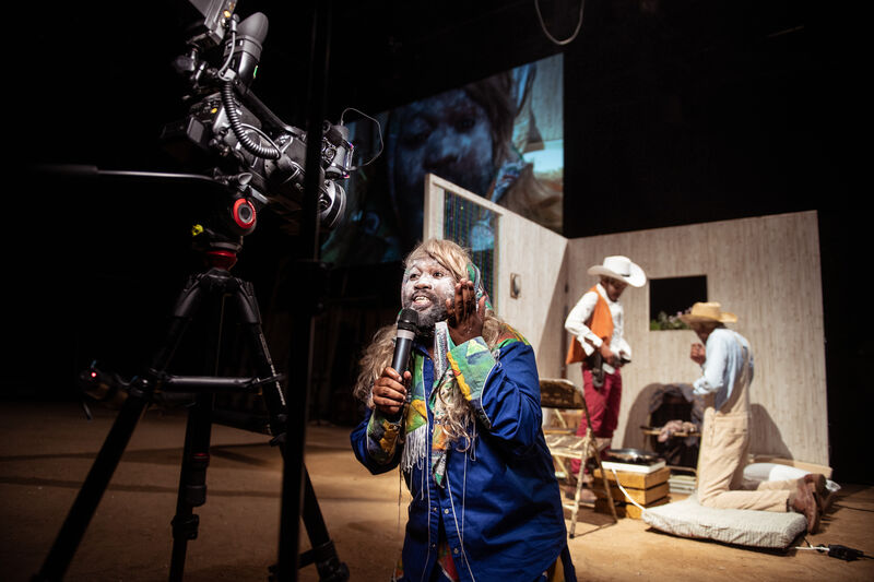 A man with a painted white face stands in front of a scene where two men are conversing dressed as cowboys and there is a camera directly in his face