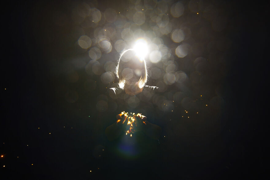 A person holds a bundle of fairylights in front of her, barely visible from a bright backlight and dust specks 