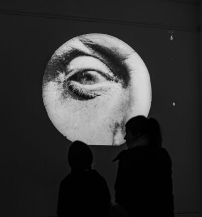Two people are stood silhouetted in front of a projection. The projection shows the close up of an eye in black and white