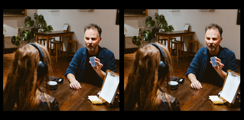 A white man with short hair sits at a table, he is holding up a blue playing card. Sitting opposite him is a girl wearing headphones