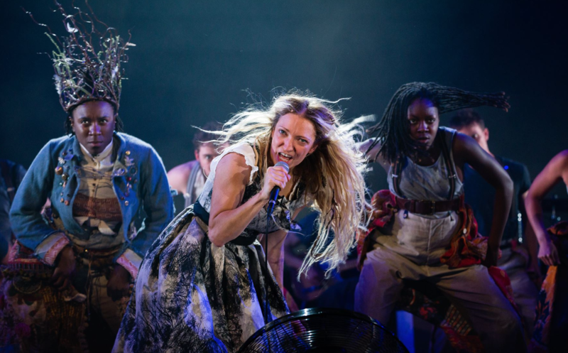 A blonde, long haired woman sings in to a microphone, behind her are two people, their arms on their thighs