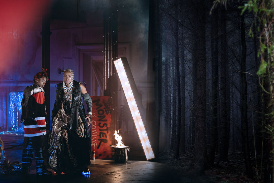 An image of the production 'Galatea' showing two performers on stage in a dystopian city environment with trees also visible