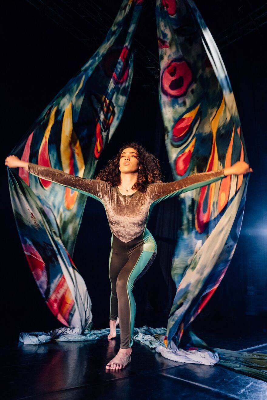 A young female performer of colour stands, arms outstretched. In her hands she hold on to two large pieces of material that are hanging from the ceiling. These look like big butterfly wings behind her.