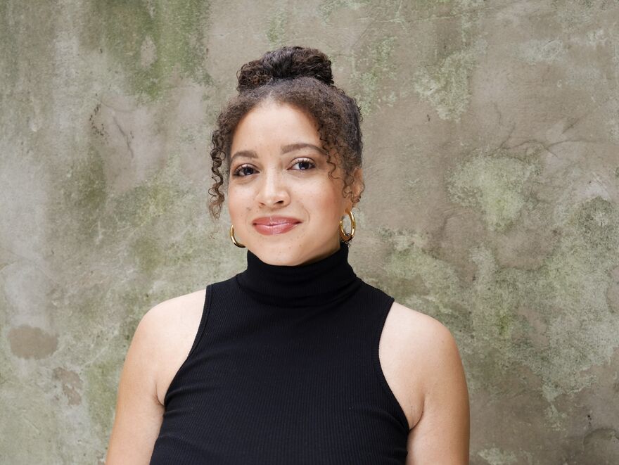 A portrait of Alexandra Sheppard. She has dark curly hair which is up in a bun and gold hoop earrings. She wears a black sleeveless turtle neck and is stood in front of a dappled background