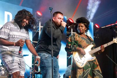From left to right a black lady with long braided her wearing white and black patterned shorts and shirt holds a microphone, a white man wearing jeans and a jacket sings into a mic in the centre, and a black lady with a dark afro wearing a patterned dress plays bass at the end. They're stood on stage performing to an audience