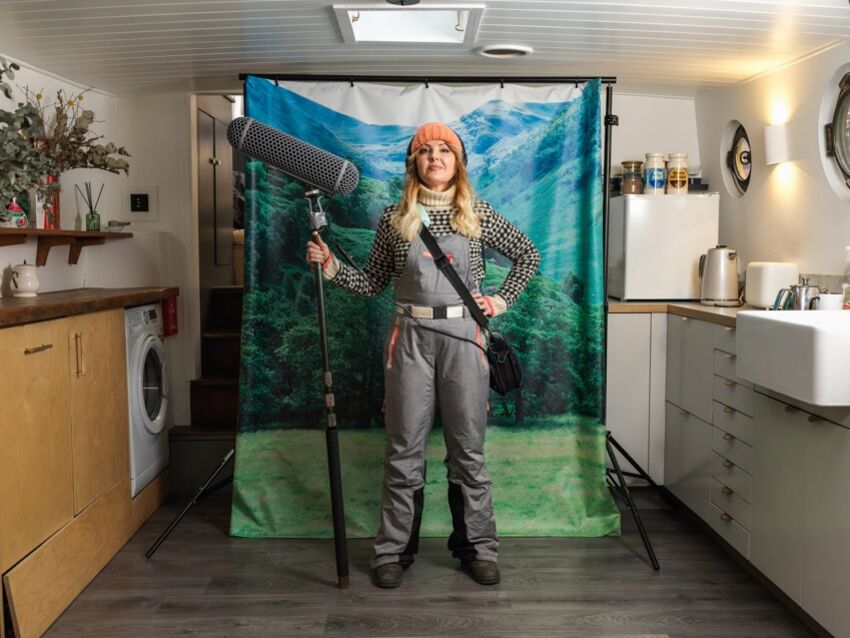 A woman standing in front of a roll out image of a nature shot, inside a kitchen. The woman is holding a large boom microphone and posing triumphantly