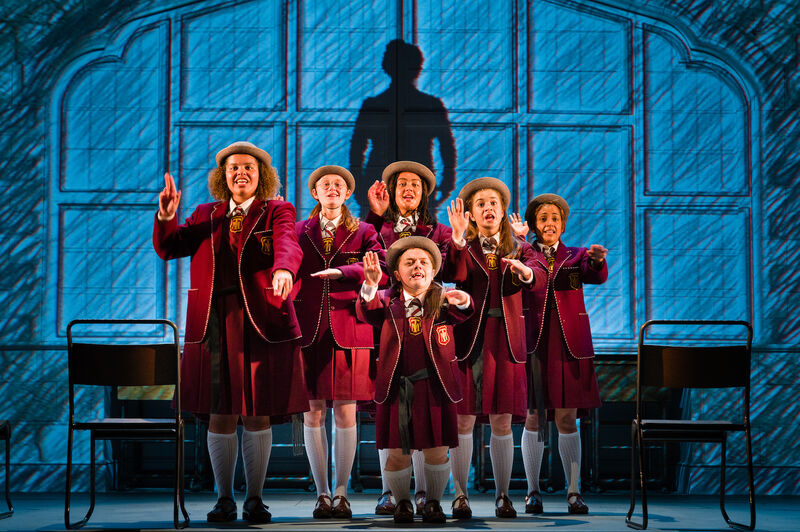 A group of girls in maroon school uniform stand in a classroom, holding one hand up and the other pointing outward