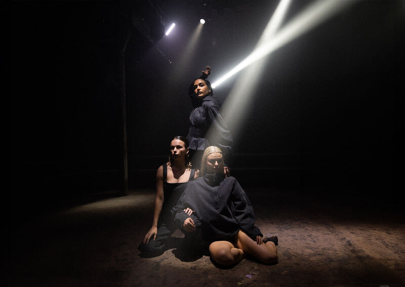 Three dancers in black clothes pose in a room lit by beams of light