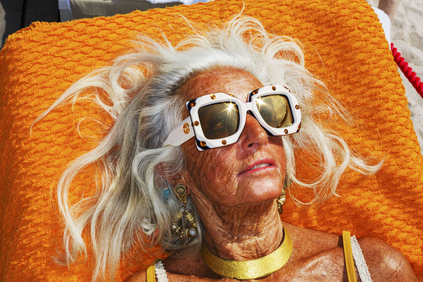 A woman with large sunglasses and gold jewelery sun bathes on an orange towel