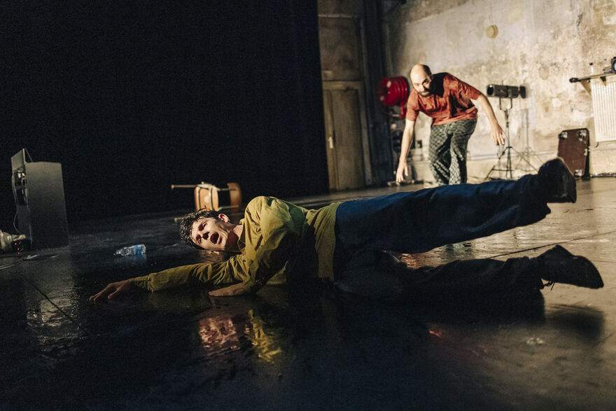 On stage picture of two performers, one is lying on the ground in the foreground and the other is bending down to look at him in the background.