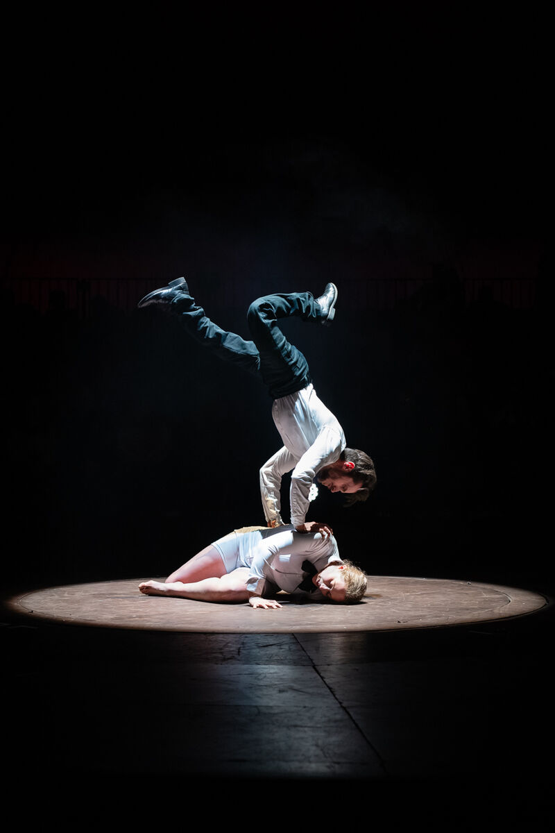 A woman lay on her side in a spotlight whilst a man does a handstand on top of her