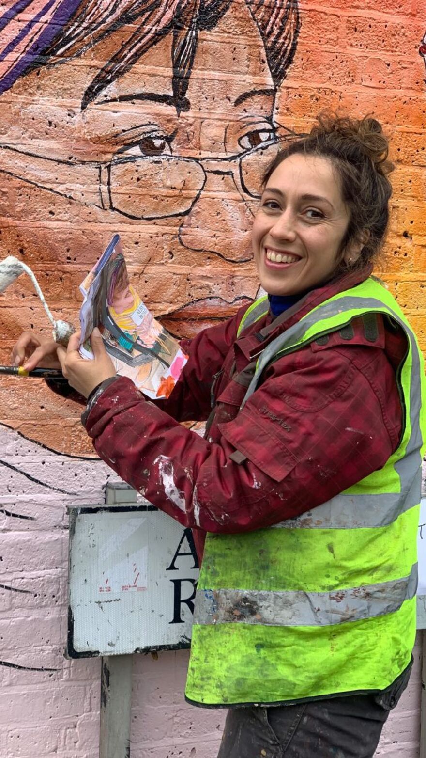 An artist in a high-vis jacket stands in front of a mural she is painting. She has a roller and a paint brush in her hands.