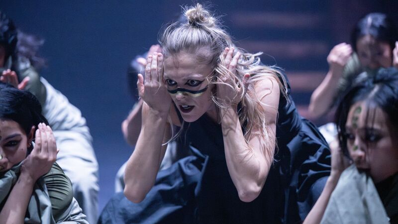 A woman with blonde hair is bent over staring intently in front of her. Her hands are held beside her face and she has stripes painted across her cheeks