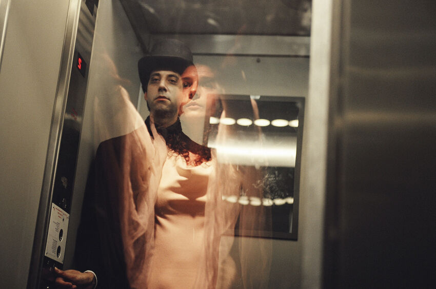 A person in a black jacket and black top hat stands in a lift, there is a ghostly figure of a person in white over the top