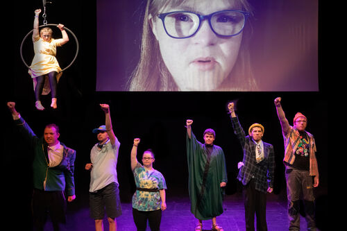 On stage. A screen behind with a woman's face wearing blue glasses and blonde hair with a fringe. Six performers on stage wearing different 'everyday' clothing with their right fists in the air. A woman in a yellow dress is on a hanging circus hoop above left of them