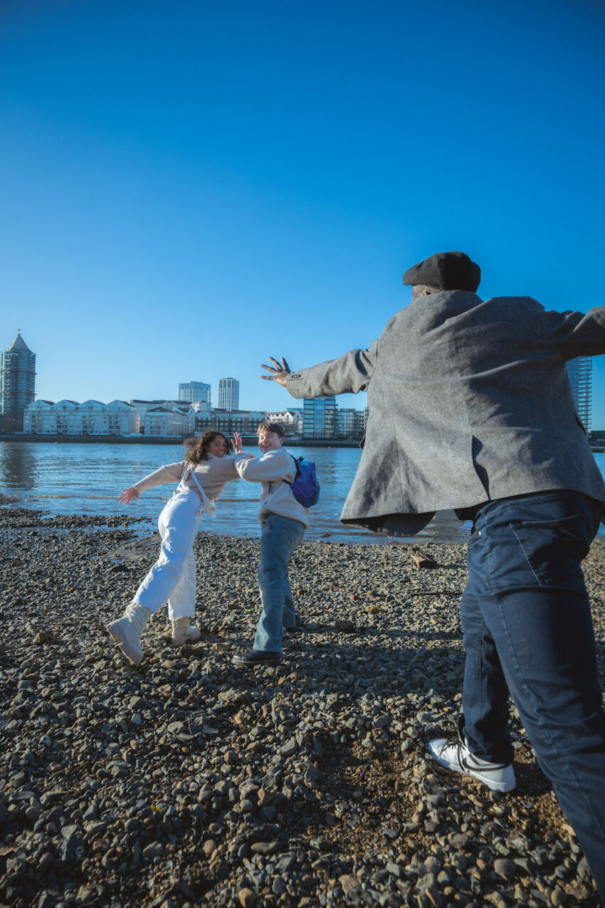 Three dancers stand on a pebble bank of a river with a city visible on the other bank in the background. Two of the dancers look back towards the camera laughing and pretending to run away, while the third dancer reaches after them
