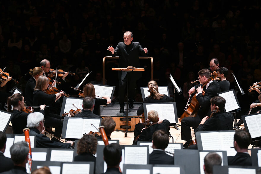 François-Xavier Roth stands poised, conducting the London Symphony Orchestra. He is central and facing the camera with the orchestra in front of him, backs to the camera, mid-performance.