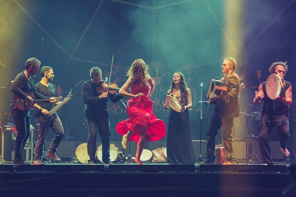 A person in a red dress dances, centre stage, surrounded by musicians