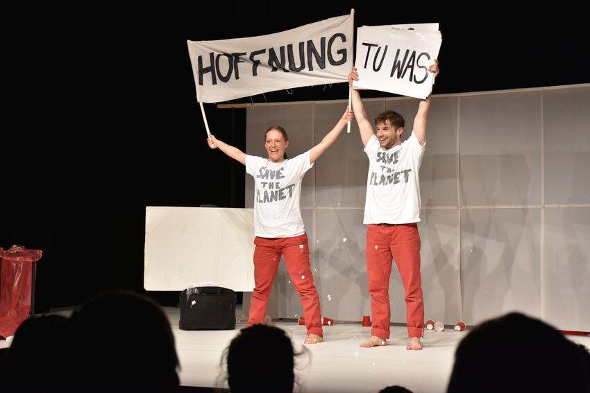 Two performers in white t-shirts and red trousers hold banners above their heads