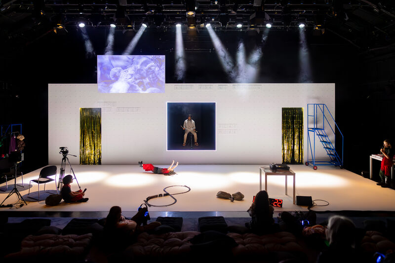 A wide shot of the stage set shows a child lying on their back with arms outstretched to the ceiling. There's a table on the right and camera equipment to the left