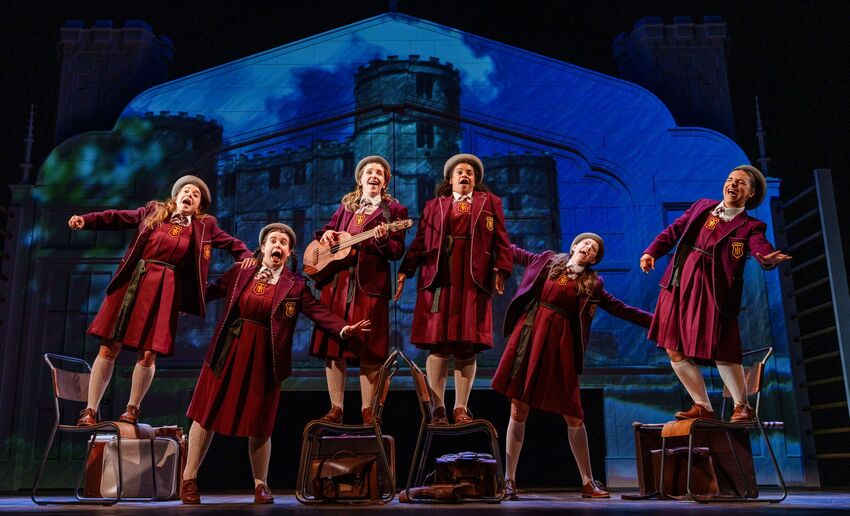 Six people in burgundy school uniforms on stage. Some are standing on chairs. One is holding a guitar and they all look like they're sining.
