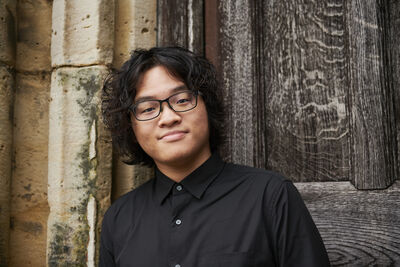Curtis Phill Hsu wearing a black button shirt and glasses, he is smiling at the camera standing in front of a wooden door