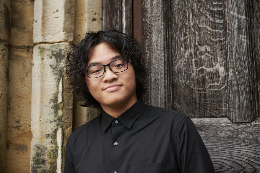 Curtis Phill Hsu wearing a black button shirt and glasses, he is smiling at the camera standing in front of a wooden door