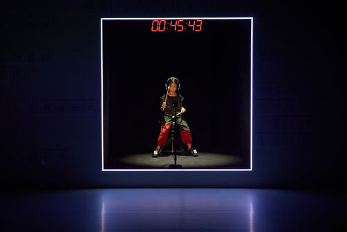 A young girl sits in darkness lit by a fluorescent frame playing a motion capture drum with a digital countdown above her