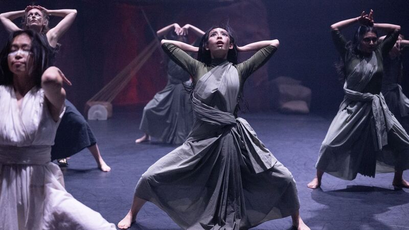 Female dancers in grey dress stand with arms above their heads and their legs spread apart, bent at the knees