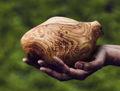 A hand, muddy from the earth, holds a carved, wooden heart that says Tender Exchange on it