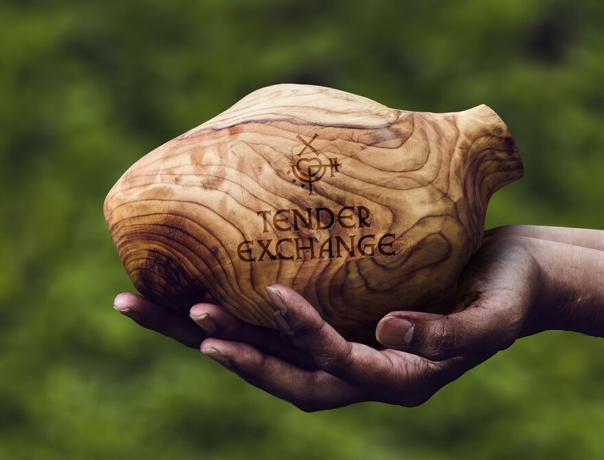 A hand, muddy from the earth, holds a carved, wooden heart that says Tender Exchange on it