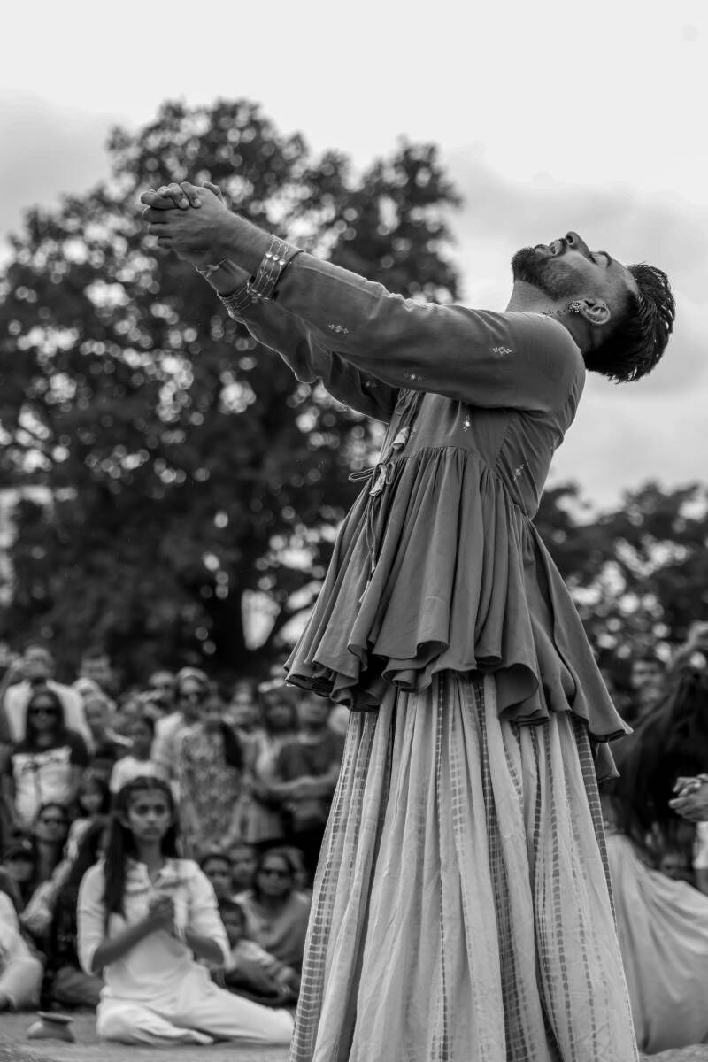 A dancer leans back, clasping his hands out in front, wearing a floaty top and skirt, audiences stood behind 