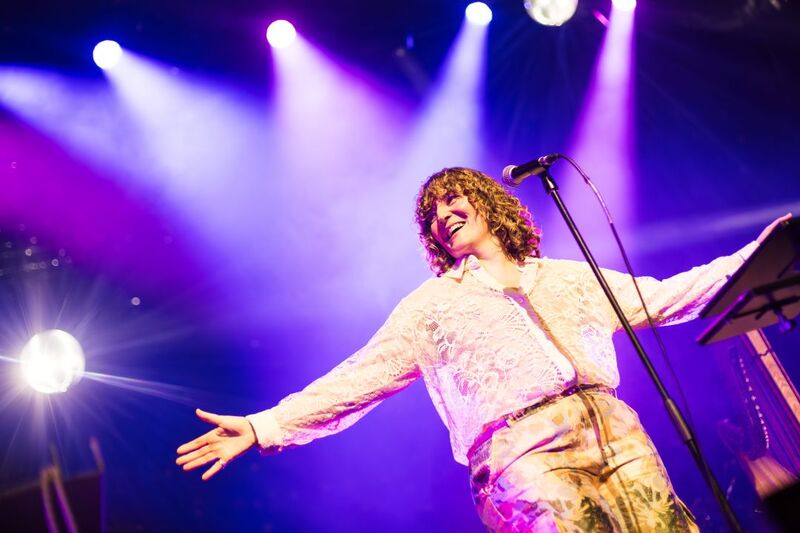 On-stage portrait of a performer standing in front of a mic with their arms raised either side of their body and smiling