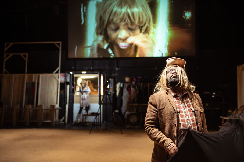 A man wearing a plaid shirt, tan jacket and blonde wig stands in front of a projection screen and stares upwards