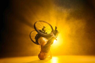 Back-lit by a bright yellow light, a man sat in a wheelchair is on his head, his wheelchair wheels raised in the air