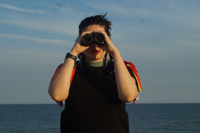 Tilly Ingram faces the camera. She is holding binoculars up to her eyes, and in the background there is the sea and the sky 